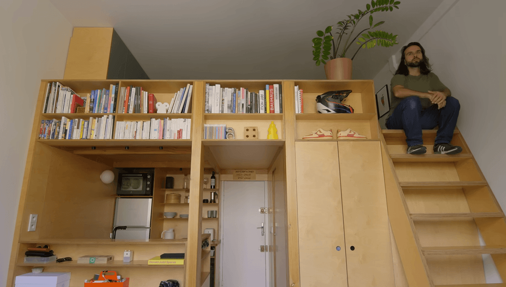 Man sits on wooden stairs next to a tall bookshelf filled with books and decor.